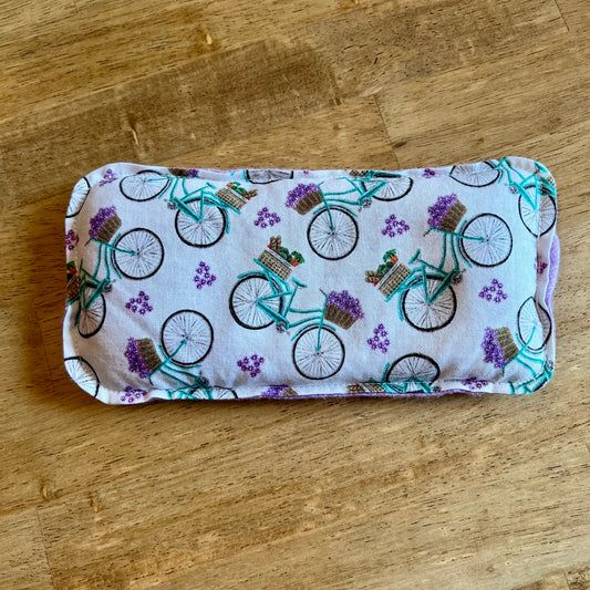 A dual-sided Petite Provence Eye Pillow by Eclectic Spruce. This pillow features a repeating pattern of teal bicycles with flower baskets on a white background.