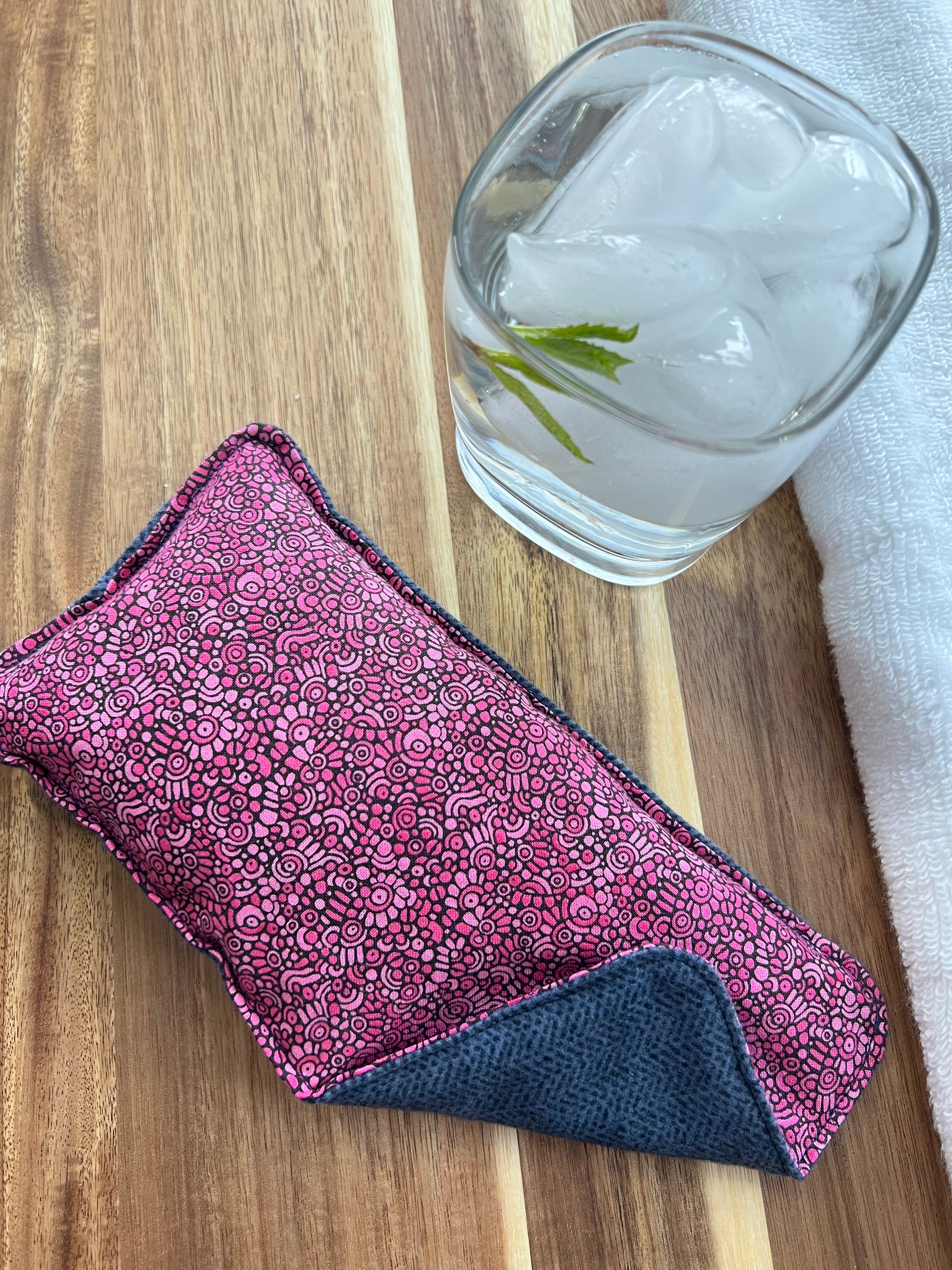 Weighted Eye Pillow on a wood surface next to a white towel and glass of ice water.