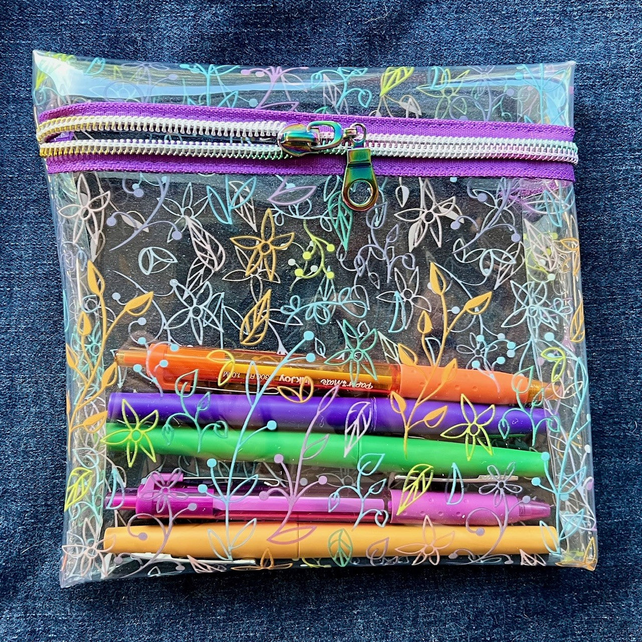 Clear see-through vinyl zipper pouch with floral pattern containing colorful pens on a blue denim background.