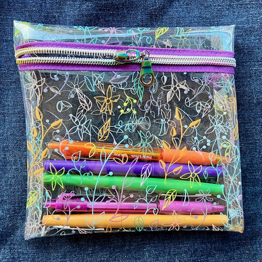 Clear Eclectic Spruce see-through vinyl zipper pouch with floral pattern containing colorful pens on a blue denim background.