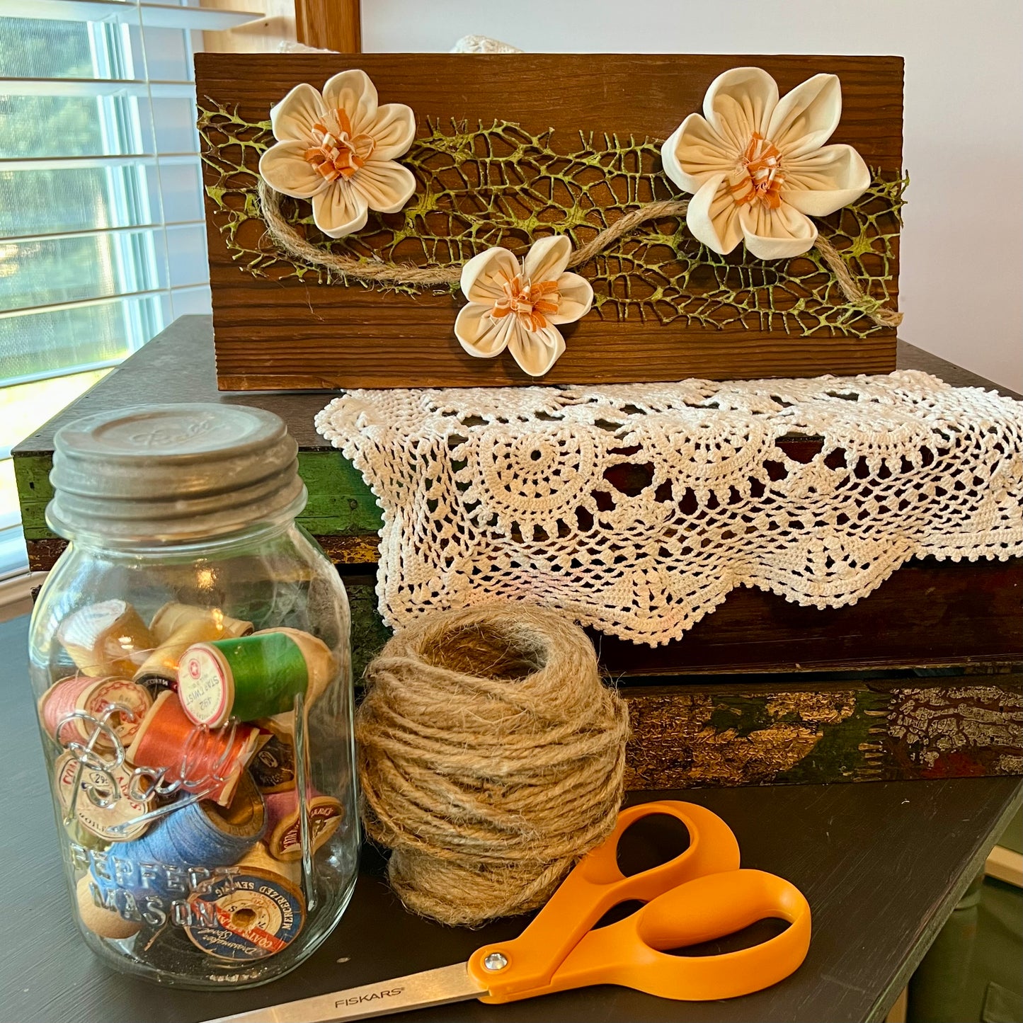 3D botanical wall art featuring cream-colored flowers made from reclaimed fabric and mounted on a dark wood plank. Displayed on a lace fabric next to a jar filled with spools of thread, a bundle of twine, and a pair of scissors.