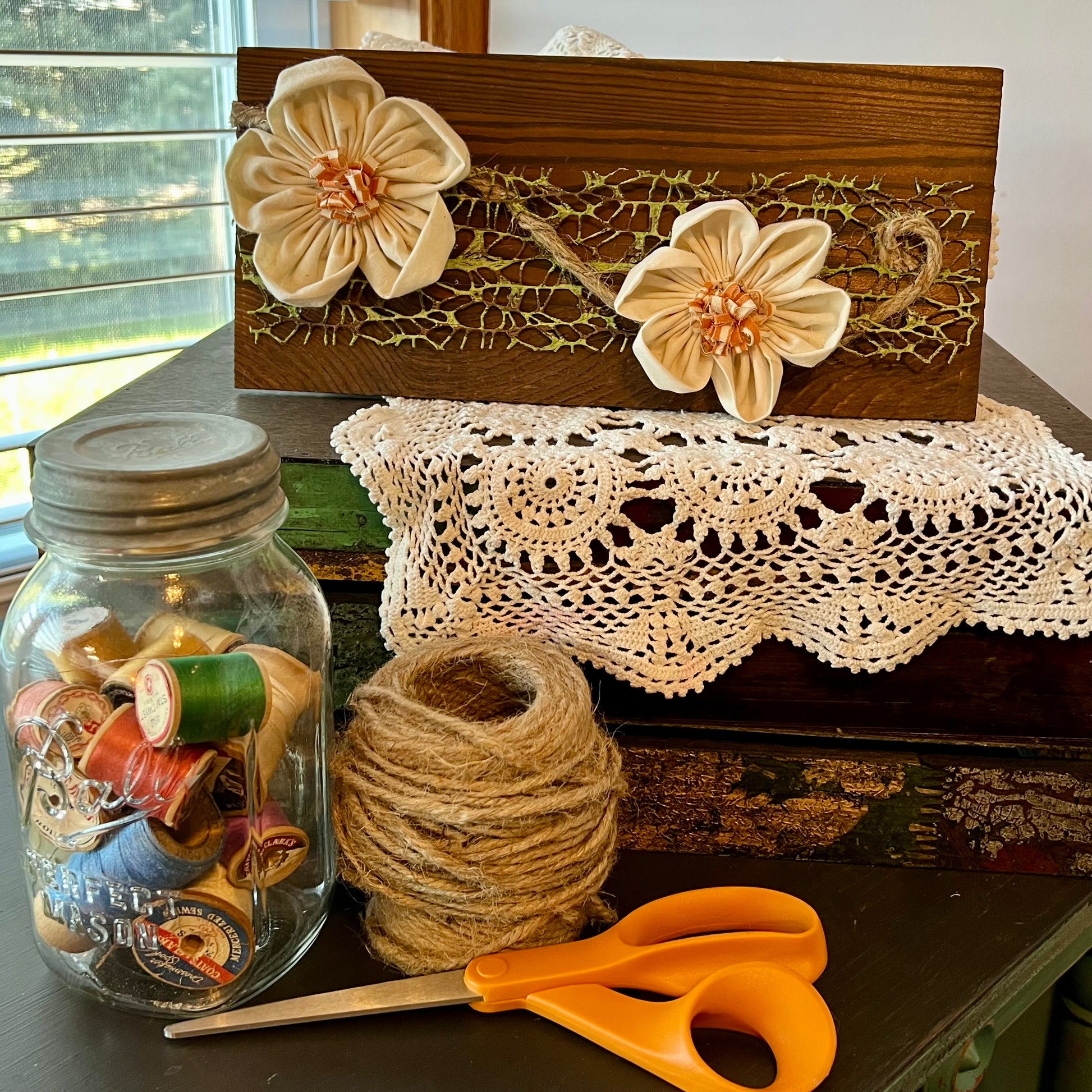 3D botanical wall art featuring cream-colored flowers made from reclaimed fabric and mounted on a dark wood plank. Displayed on a lace fabric next to a jar filled with spools of thread, a bundle of twine, and a pair of scissors.