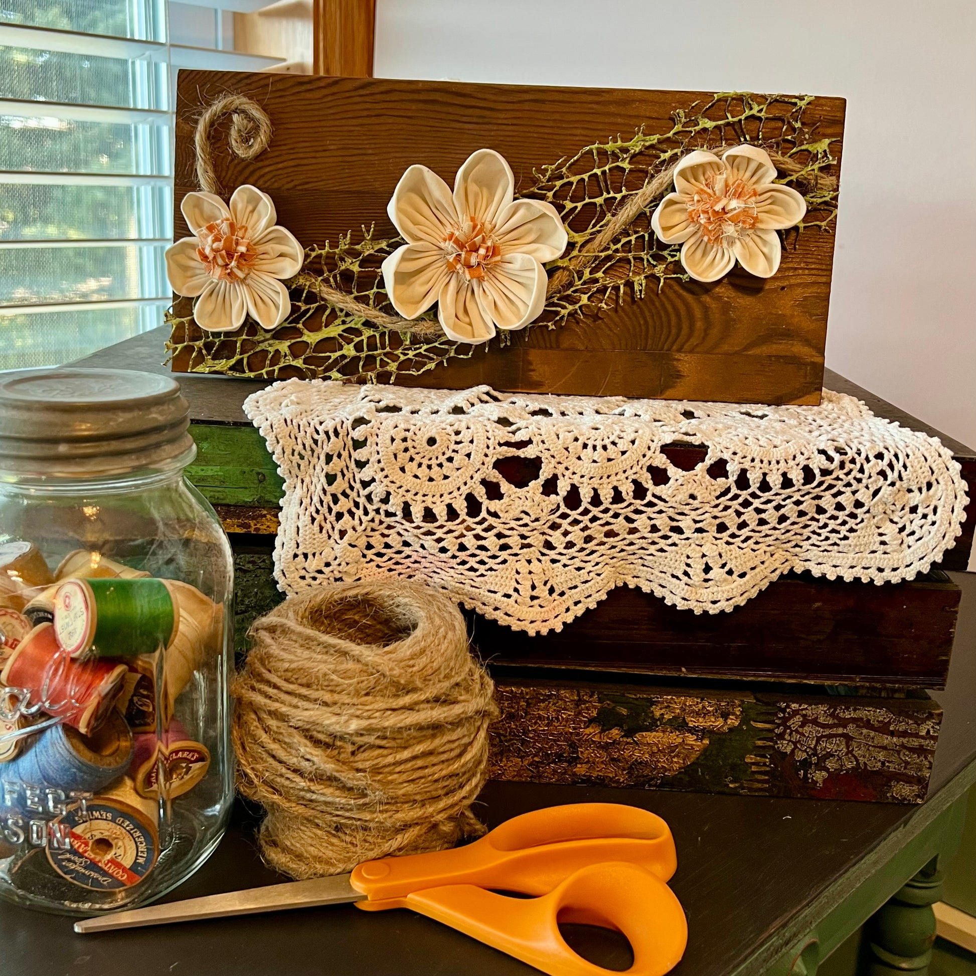 3D botanical wall art featuring cream-colored flowers made from reclaimed fabric and mounted on a dark wood plank. Displayed on a lace fabric next to a jar filled with spools of thread, a bundle of twine, and a pair of scissors.