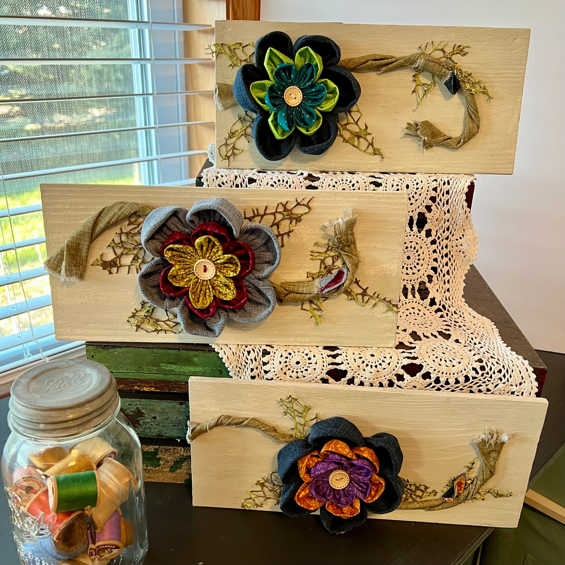 The entire Meadow Bloom collection: Three pieces of 3D botanical wall art featuring reclaimed denim and jewel-tone flowers on a distressed and painted wood plank. Displayed on three levels among lace fabric next to a jar filled with spools of thread.