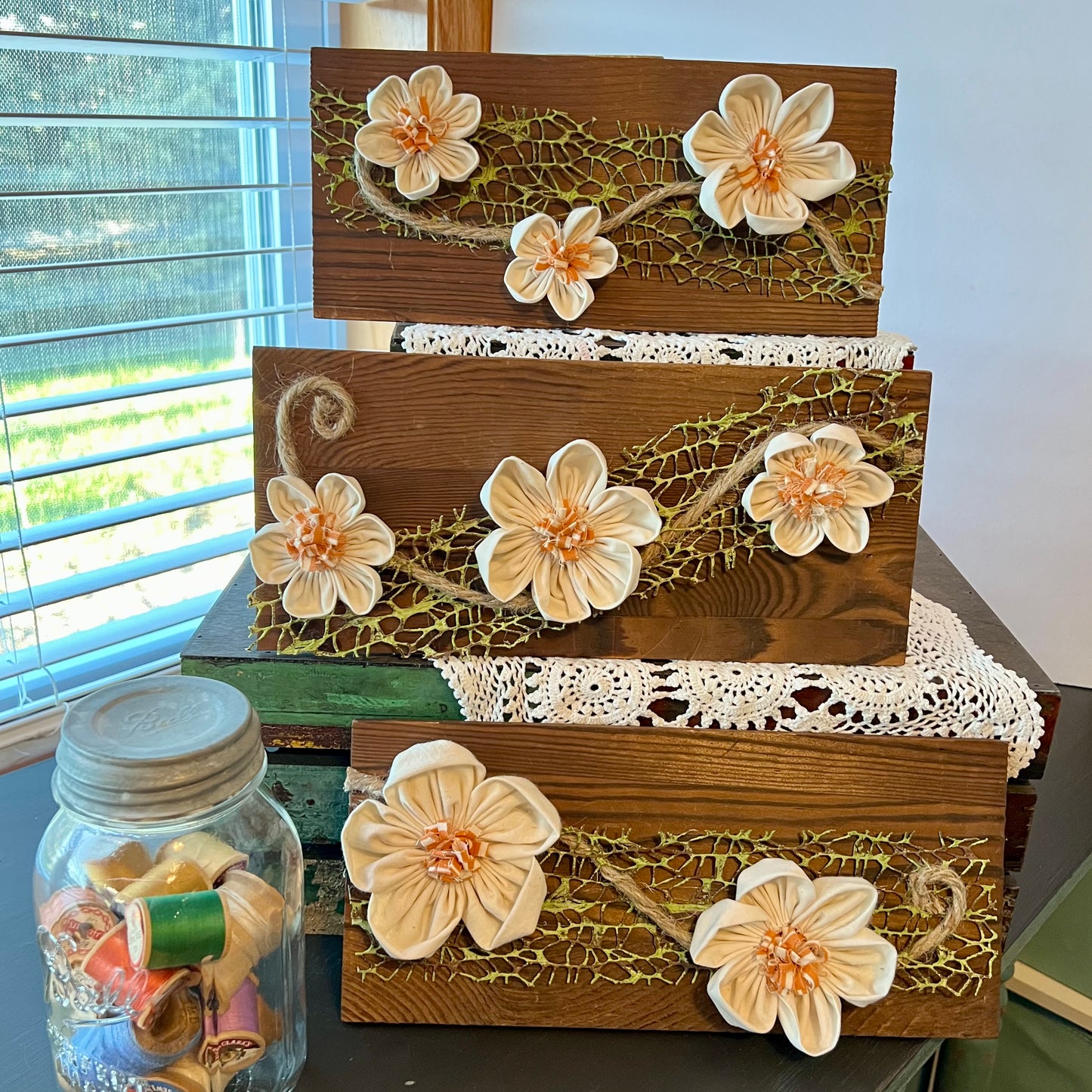 The entire Botanical Trellis collection: Three pieces of 3D botanical wall art featuring cream-colored flowers made from reclaimed fabric and mounted on a dark wood plank. Displayed on three levels among lace fabric next to a jar filled with spools of thread.