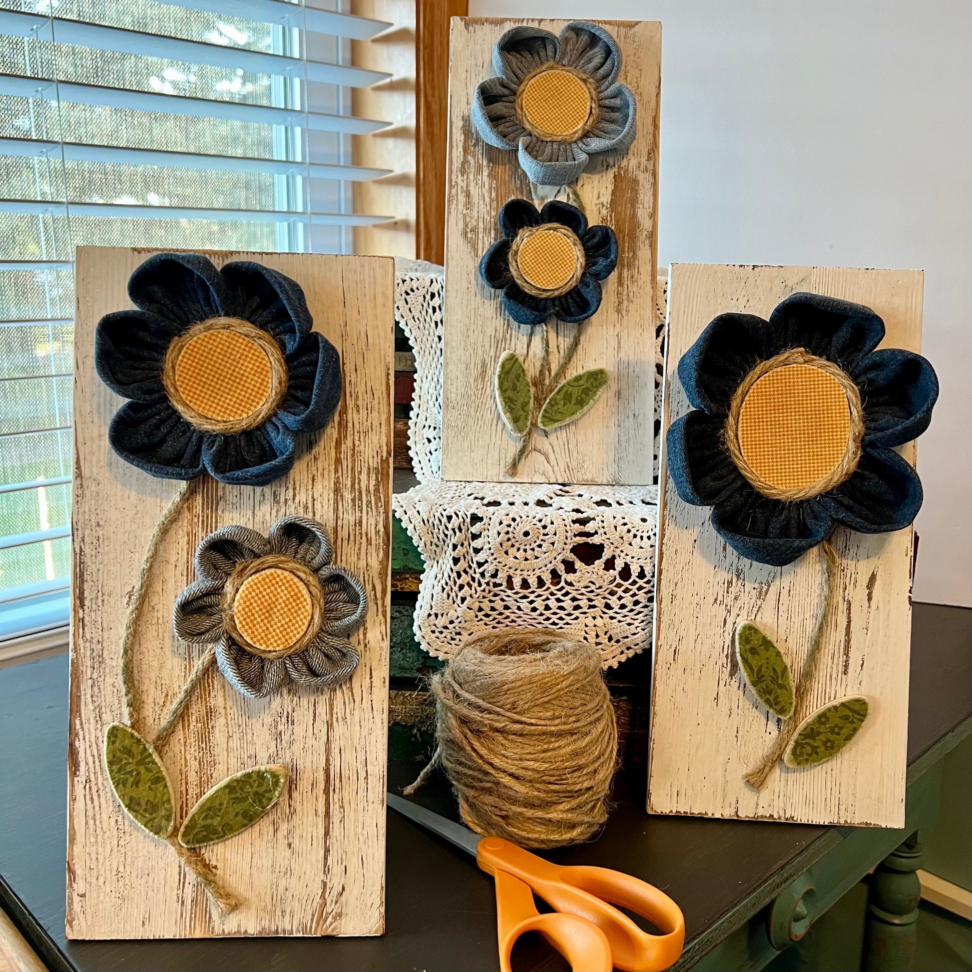 The entire Sunny Day Reclaimed Denim collection: 3D botanical wall art featuring yellow gingham and reclaimed denim flowers on a white-washed wood plank. Displayed on a lace fabric next to a bundle of twine and a pair of scissors.