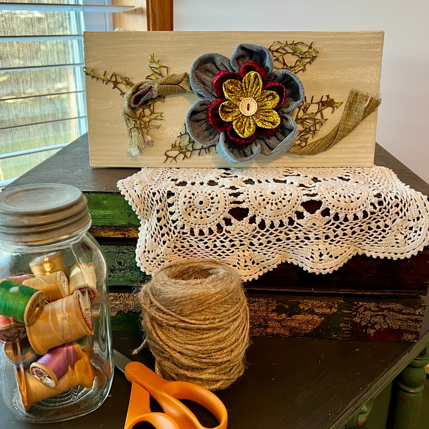 3D botanical wall art featuring reclaimed denim and jewel-tone flowers on a distressed and painted wood plank. Displayed on a lace fabric next to a jar filled with spools of thread, a bundle of twine, and a pair of scissors.