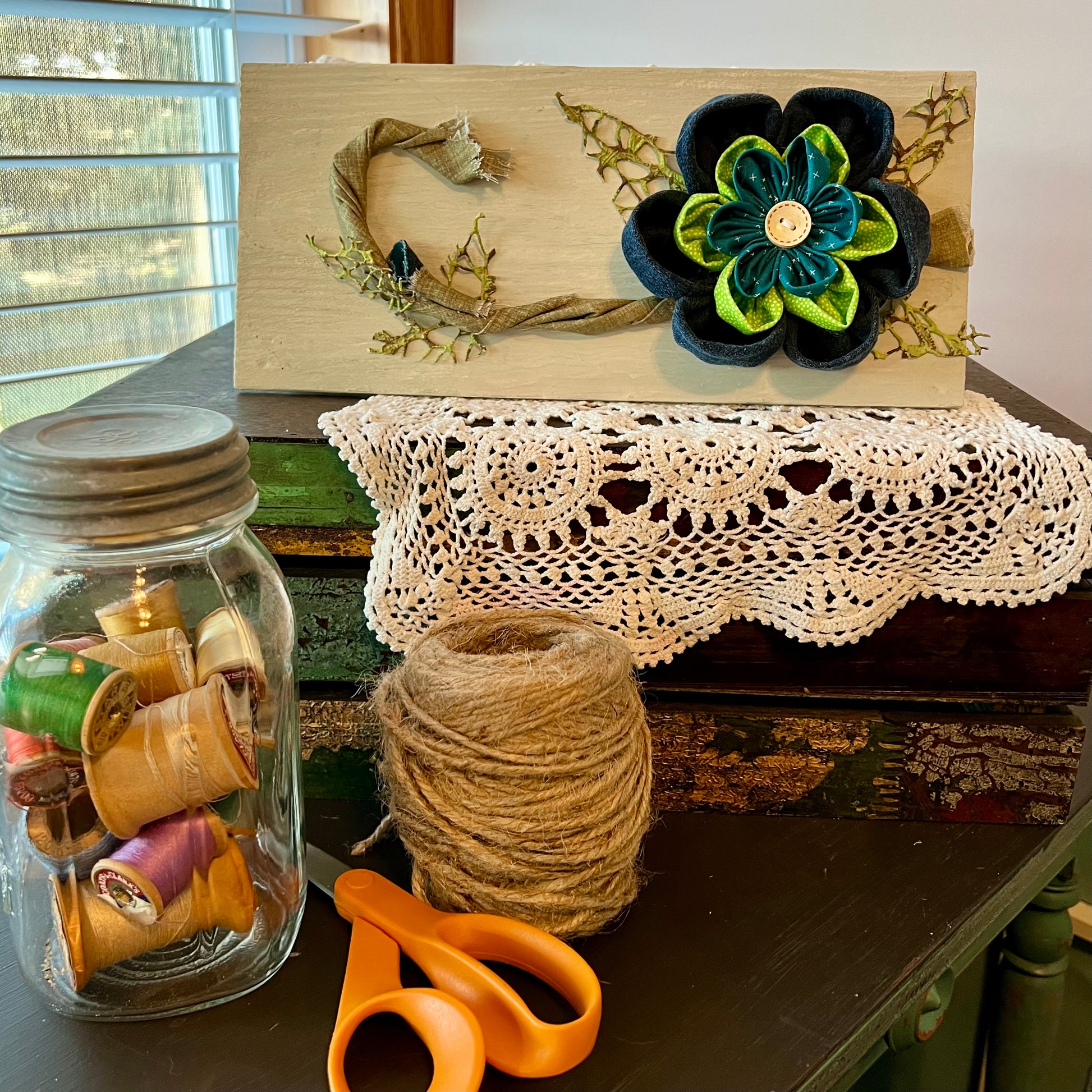 3D botanical wall art featuring reclaimed denim and jewel-tone flowers on a distressed and painted wood plank. Displayed on a lace fabric next to a jar filled with spools of thread, a bundle of twine, and a pair of scissors.
