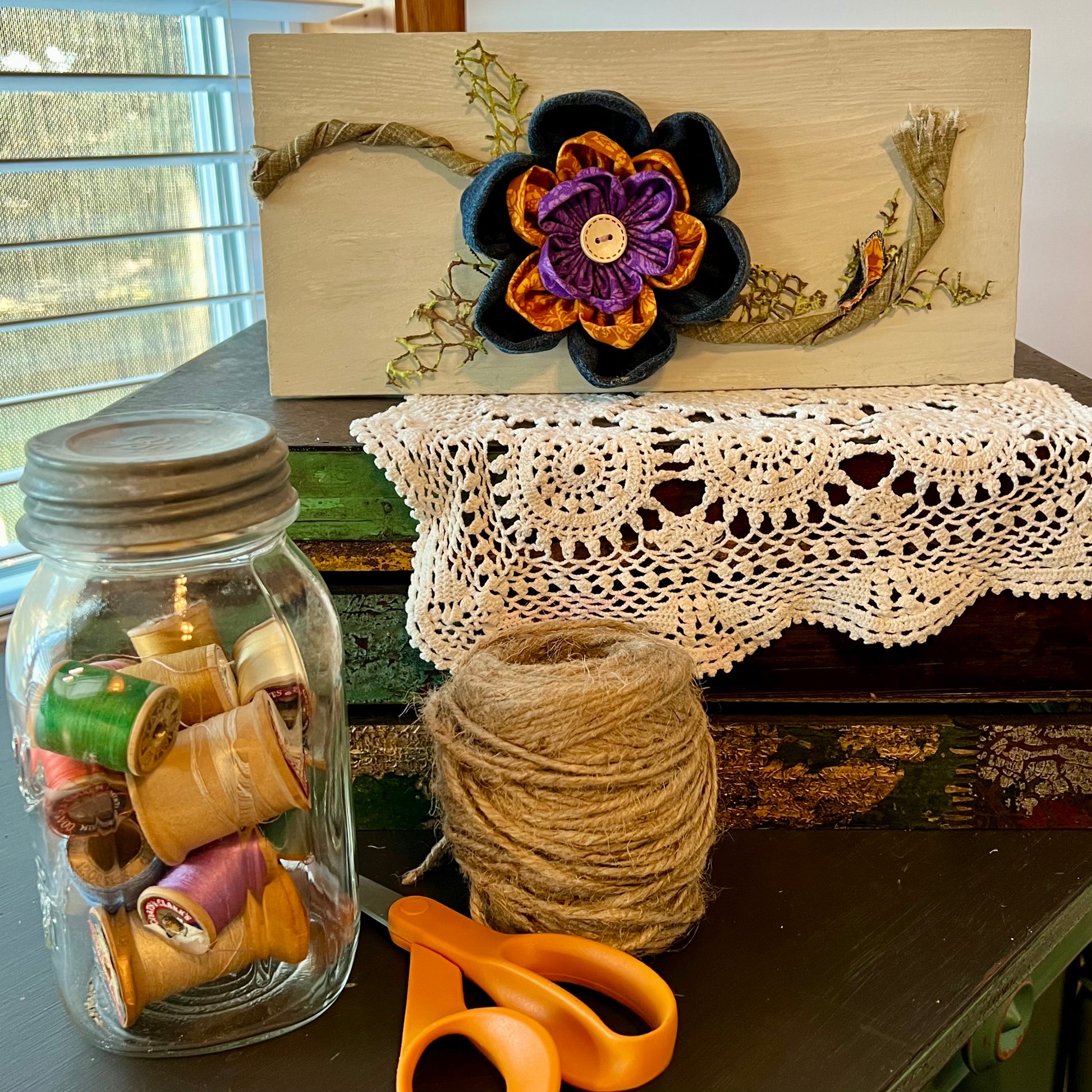 3D botanical wall art featuring reclaimed denim and jewel-tone flowers on a distressed and painted wood plank. Displayed on a lace fabric next to a jar filled with spools of thread, a bundle of twine, and a pair of scissors.