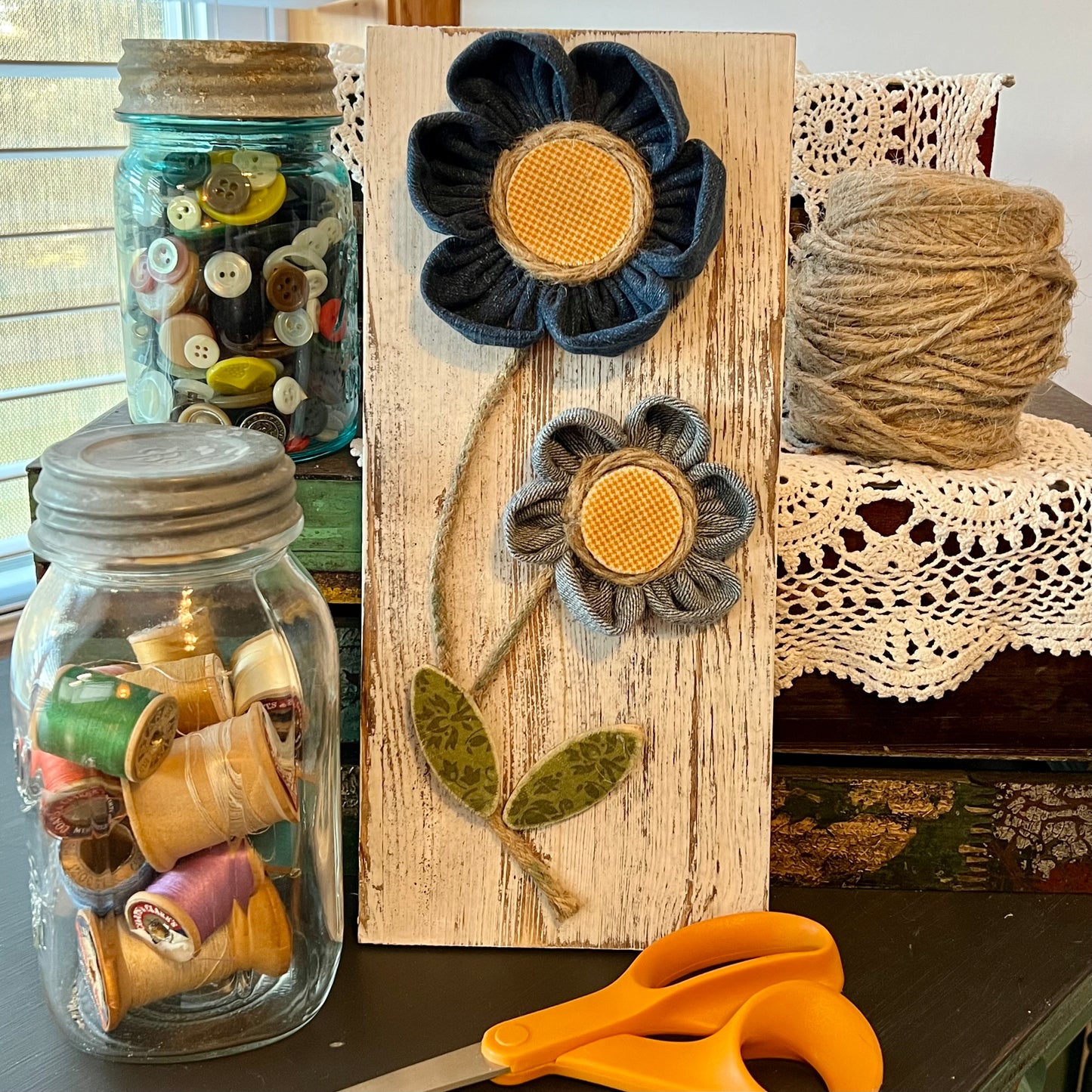 3D botanical wall art featuring yellow gingham and reclaimed denim flowers on a white-washed wood plank. Displayed on a lace fabric next to a jar filled with spools of thread, a bundle of twine, a jar of buttons, and a pair of scissors.