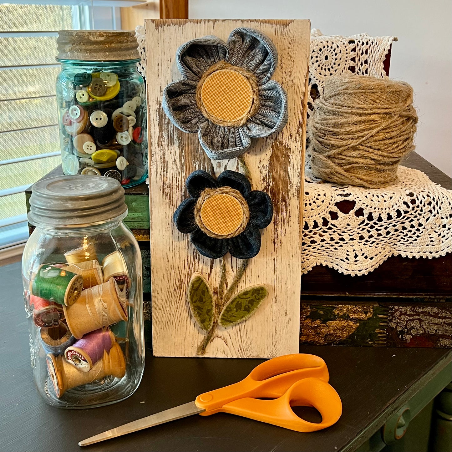 3D botanical wall art featuring yellow gingham and reclaimed denim flowers on a white-washed wood plank. Displayed on a lace fabric next to a jar filled with spools of thread, a bundle of twine, a jar of buttons, and a pair of scissors.