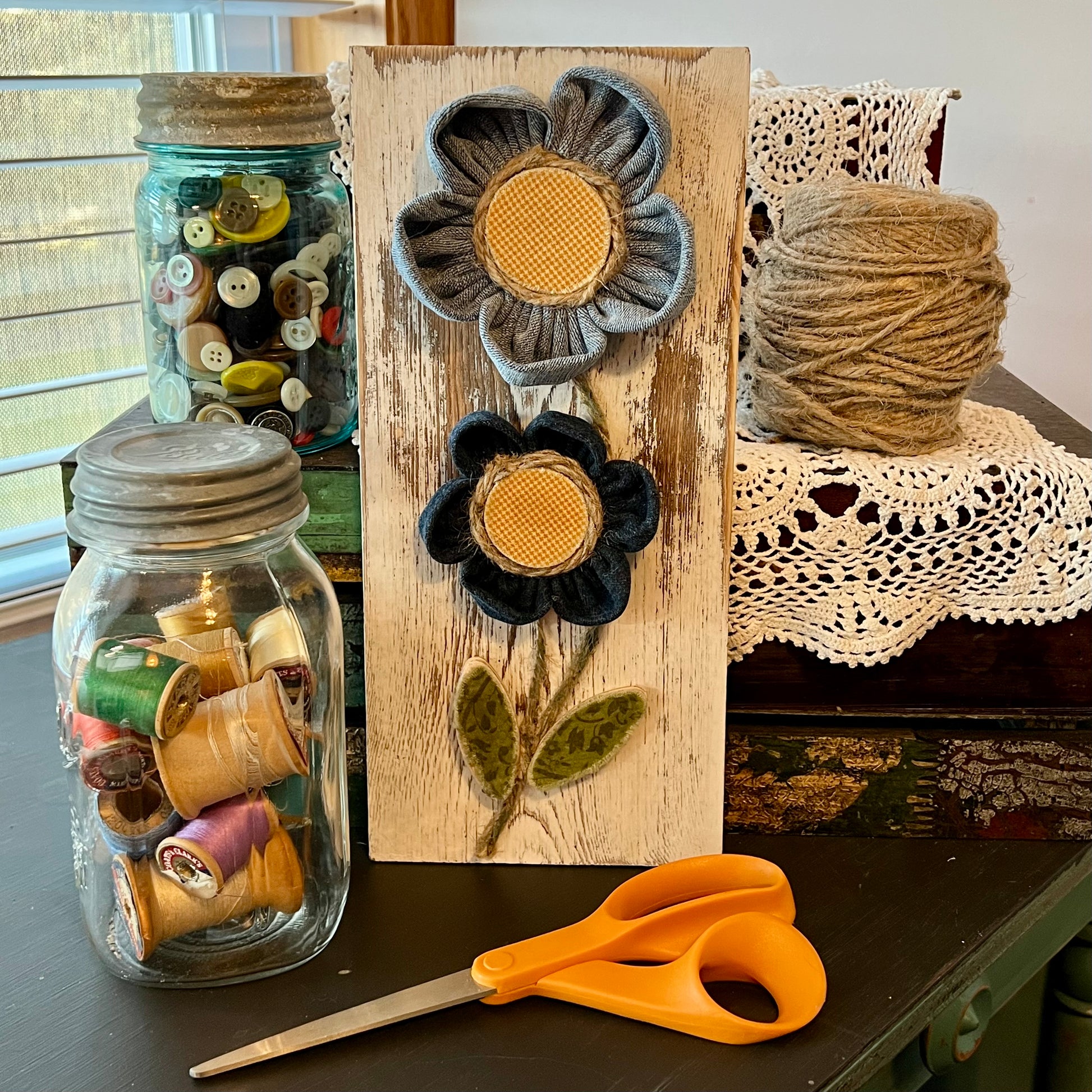 3D botanical wall art featuring yellow gingham and reclaimed denim flowers on a white-washed wood plank. Displayed on a lace fabric next to a jar filled with spools of thread, a bundle of twine, a jar of buttons, and a pair of scissors.