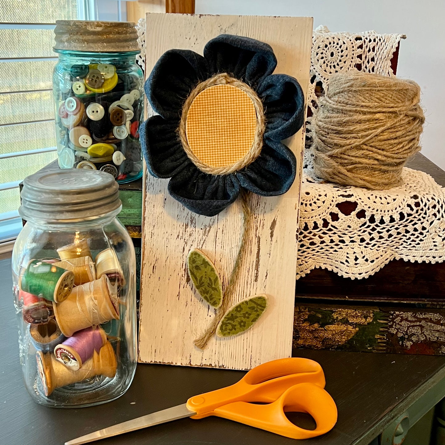 3D botanical wall art featuring yellow gingham and reclaimed denim flowers on a white-washed wood plank. Displayed on a lace fabric next to a jar filled with spools of thread, a bundle of twine, a jar of buttons, and a pair of scissors.