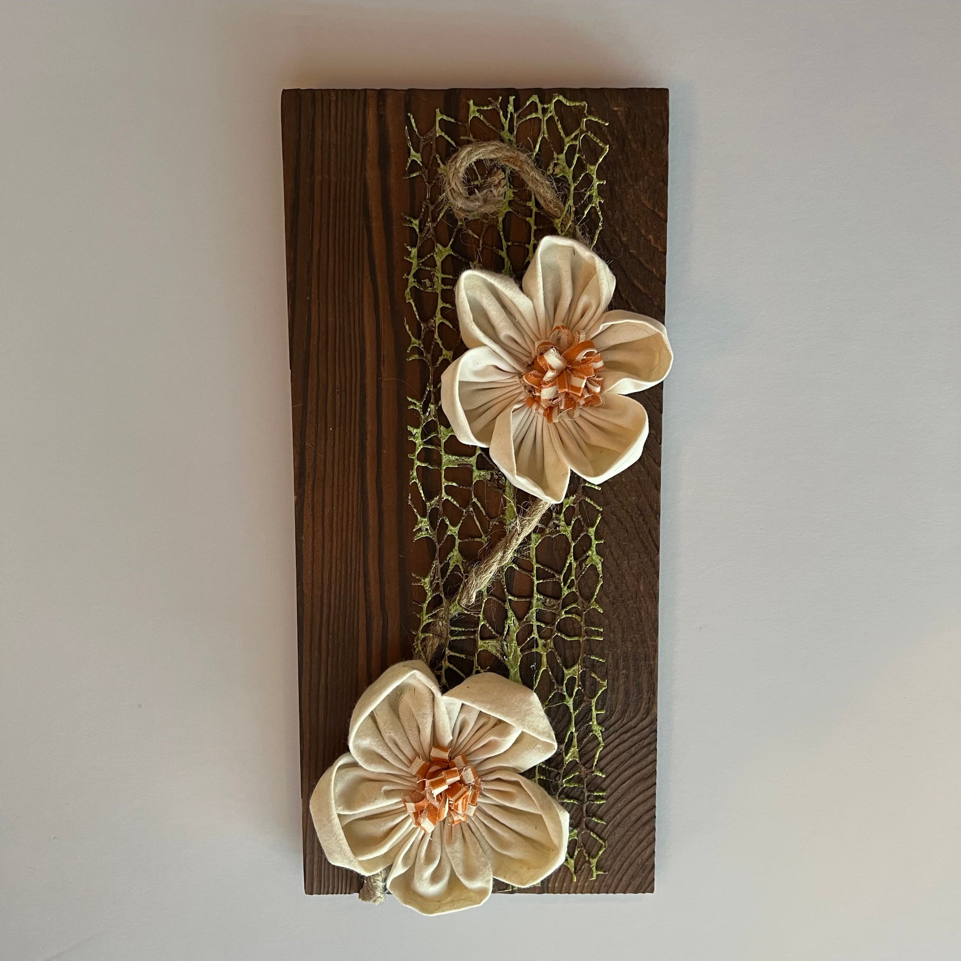 3D botanical wall art featuring cream-colored flowers made from reclaimed fabric and mounted on a dark wood plank. Displayed on a lace fabric next to a jar filled with spools of thread, a bundle of twine, and a pair of scissors.