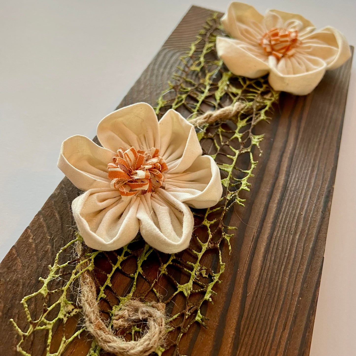 Close-up of the 3D botanical wall art featuring cream-colored flowers made from reclaimed fabric and mounted on a dark wood plank.