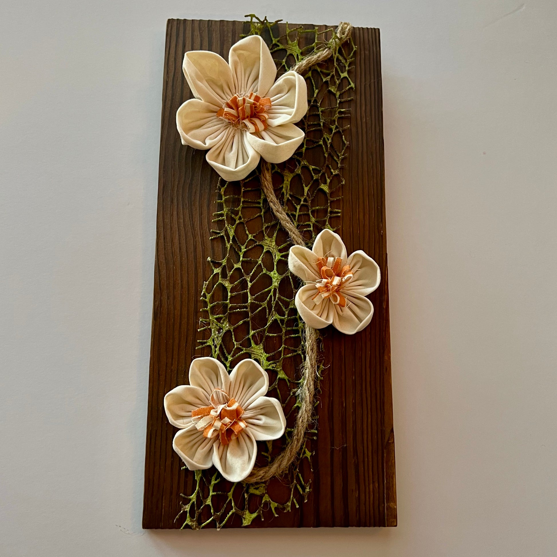 3D botanical wall art featuring cream-colored flowers made from reclaimed fabric and mounted on a dark wood plank.