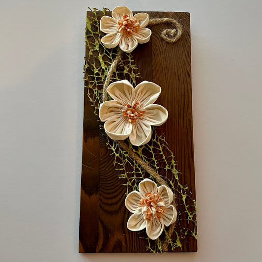 3D botanical wall art featuring cream-colored flowers made from reclaimed fabric and mounted on a dark wood plank.
