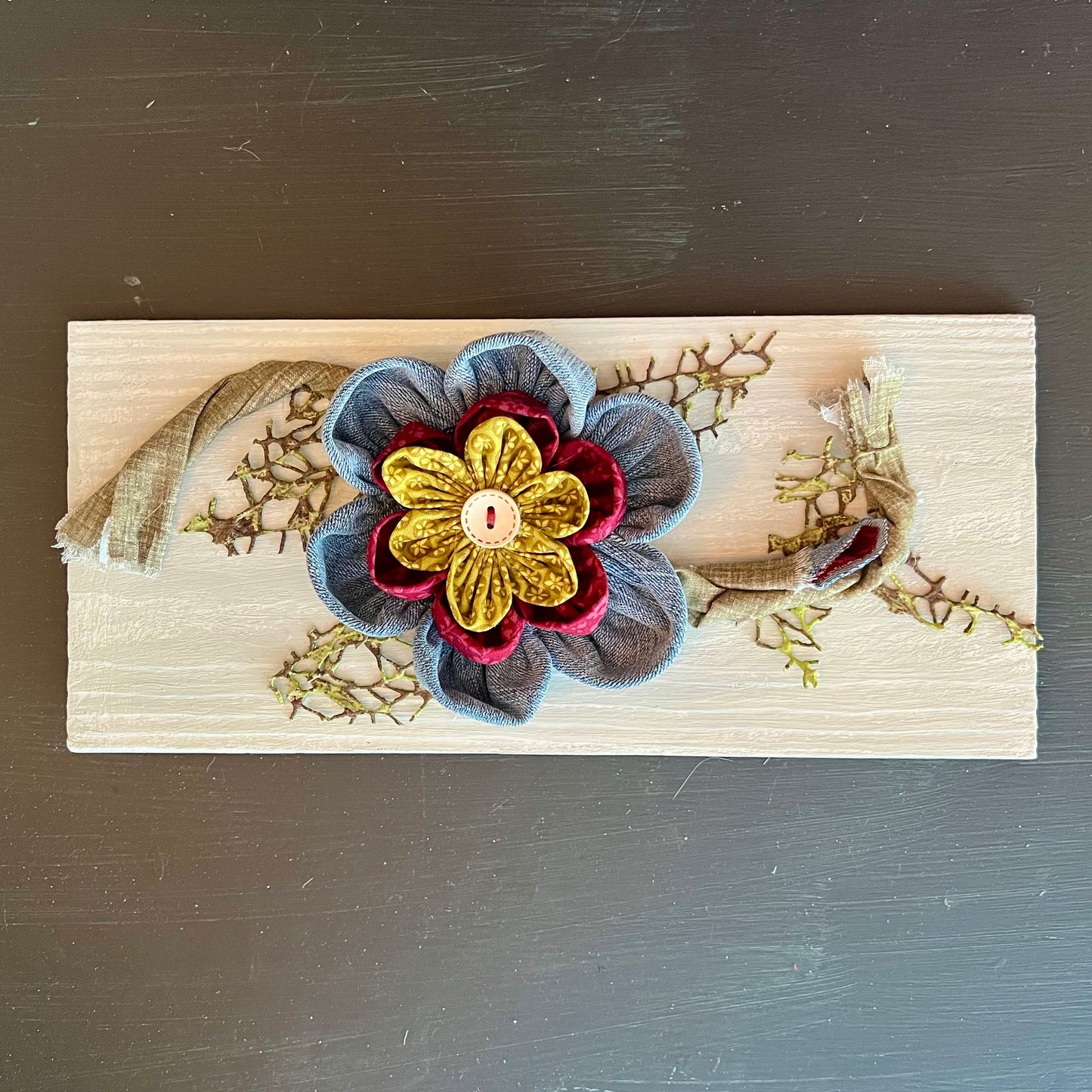 3D botanical wall art featuring reclaimed denim and jewel-tone flowers on a distressed and painted wood plank. 