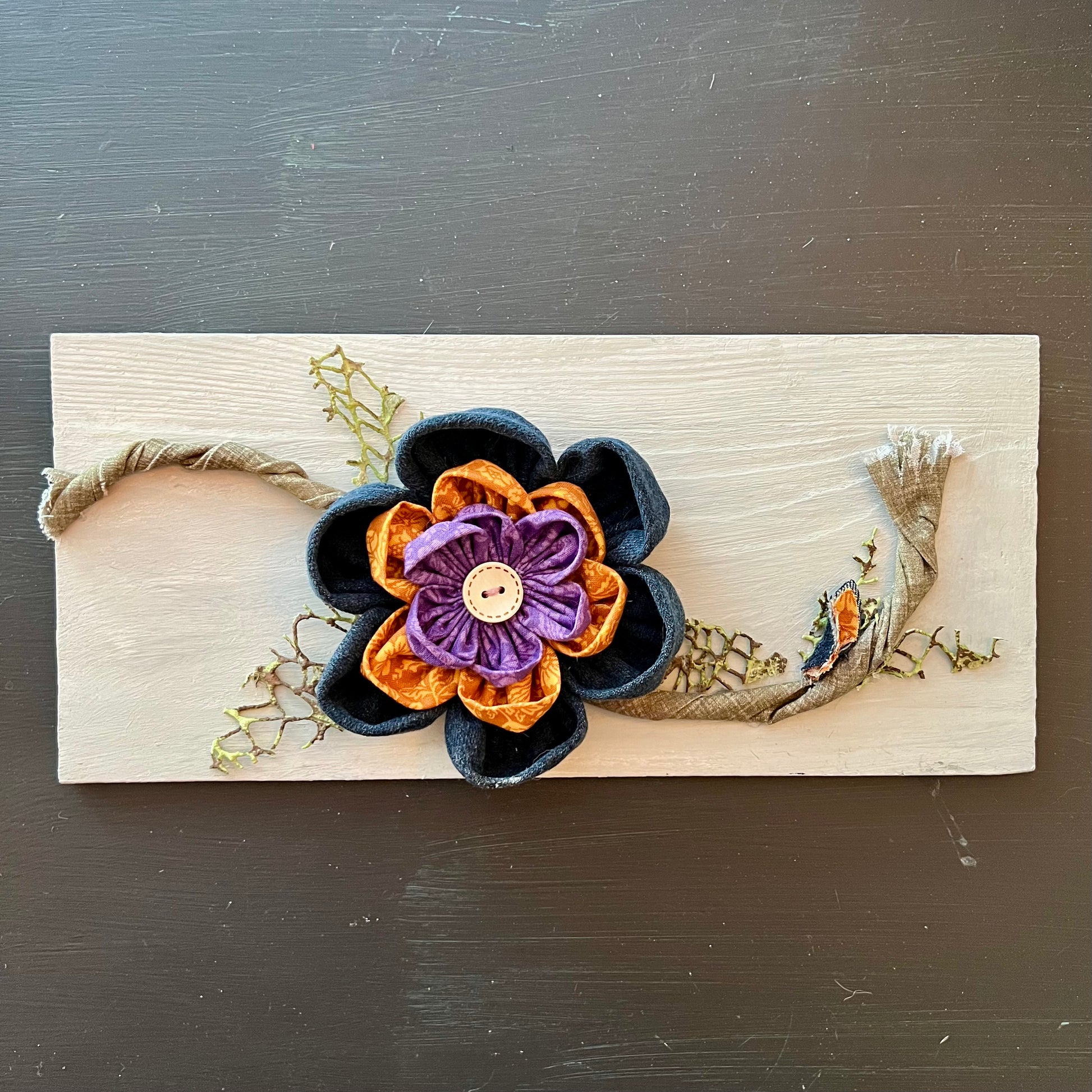 3D botanical wall art featuring reclaimed denim and jewel-tone flowers on a distressed and painted wood plank. 