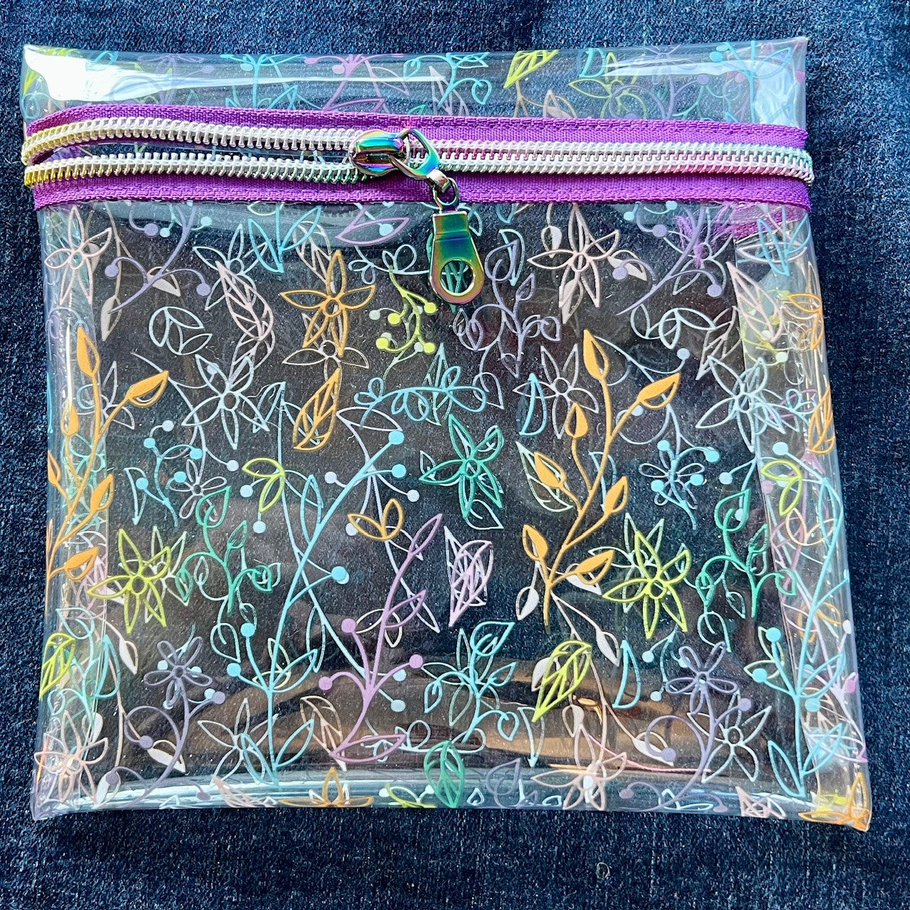 Clear see-through vinyl zipper pouch with floral pattern on a blue denim background.