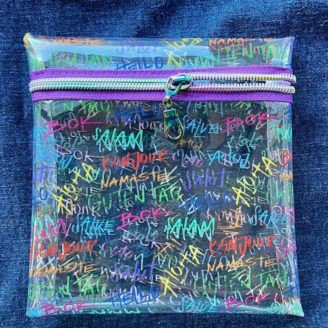 Clear see-through vinyl zipper pouch with colorful text saying "hello" in various languages and a purple zipper on a blue denim background.